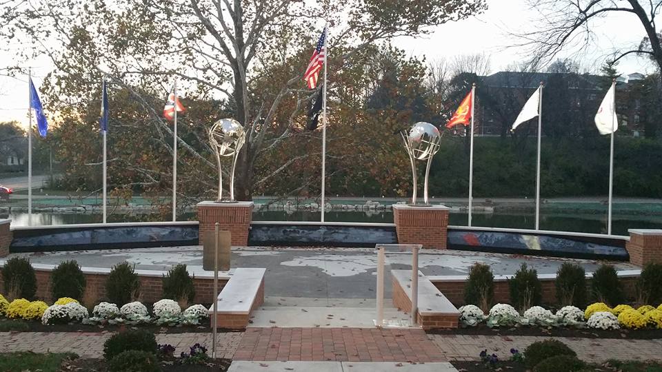 A Veterans Kiosk memorial features two globe sculptures on brick pedestals, surrounded by seven flags, including the American flag. White and yellow flowers adorn the foreground, with a tree and water providing a serene background.