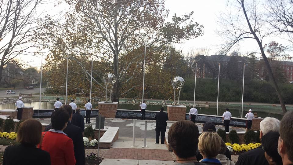 People in uniform stand in a line during a ceremony near flagpoles and a reflecting pool, surrounded by trees and brick structures. Attendees observe from a paved area, some near a veterans kiosk. Its a calm setting, possibly a memorial or official event.