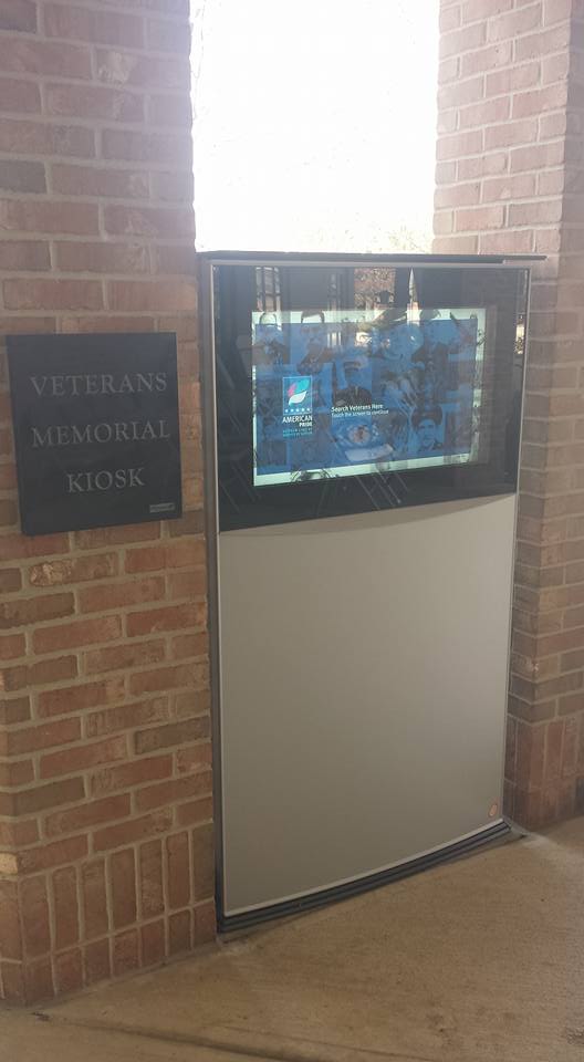 A Veterans Kiosk is mounted on a brick wall, displaying information and images honoring veterans. The digital screen enhances the memorial sites tribute atmosphere.