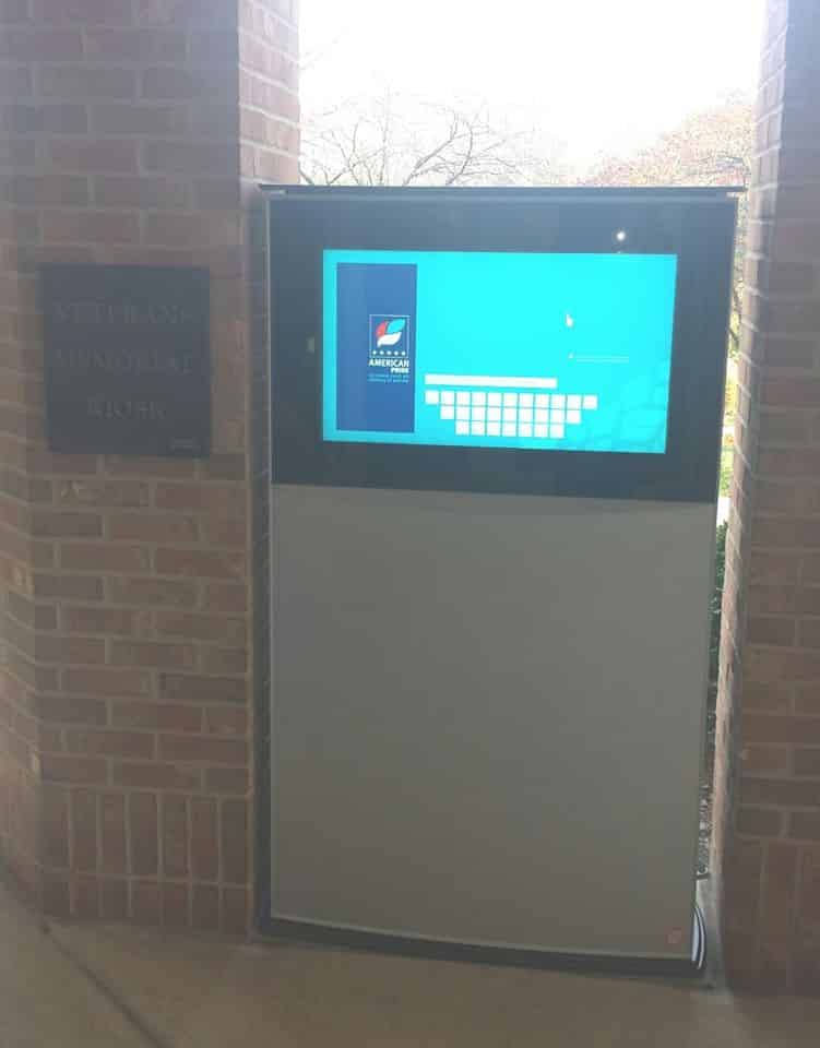 An outdoor electronic kiosk, dedicated as a Veterans Kiosk, is embedded in a brick wall. Its touchscreen display showcases a blue interface with a keyboard layout. A plaque honoring veterans is mounted beside it.