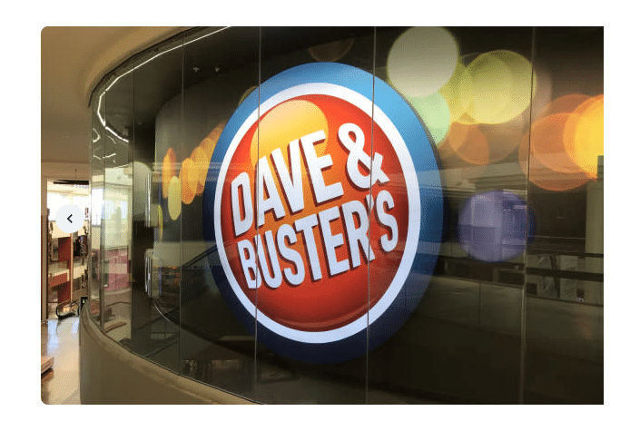 A circular sign displaying Dave & Busters is prominently featured on a glossy wall next to an interactive kiosk. The background has colorful, out-of-focus lights, giving a lively and energetic feel to the scene.