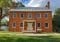 A historic red brick house with two front doors and several windows rests under a partly cloudy sky, resembling a classic case study in architecture. The metal roof and small porch with two columns complete the scene, surrounded by grass and a few trees.