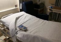 A hospital bed with white sheets and a remote control rests beside a dark blue reclining chair in a medical room, featuring a tray table, medical equipment, and Alexa Patient Self-Service for added convenience.