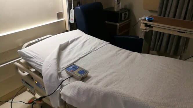 A hospital bed with white sheets and a remote control rests beside a dark blue reclining chair in a medical room, featuring a tray table, medical equipment, and Alexa Patient Self-Service for added convenience.