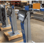 Several interactive kiosks, some with screens removed, are lined up on a factory floor. The area is spacious and organized, featuring industrial shelving in the background. Some kiosks are on wooden pallets, hinting at setup or maintenance work for digital signage installations.
