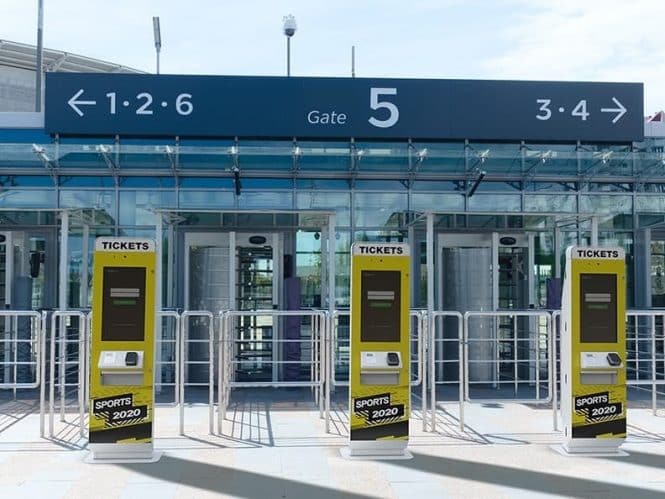 Ticket kiosks for Sports 2020 stand proudly in front of a glass building entrance at Gate 5, enhanced by sleek digital signage. Overhead signs guide visitors to gates 1-2, 3-4, and 6. The outdoor scene unfolds under clear skies.