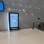 An airport terminal features digital signage with a map, guiding travelers to Gates E1 - E37. Kiosks line the path, and an escalator stands on the right. A flight information display board hovers in the upper left corner.