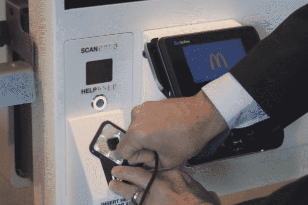 A person is attempting to charge a smartphone using a payment terminal at a McDonalds kiosk. The phone is plugged in via a cable, and the kiosk displays the McDonald’s logo on its screen.