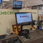 At Kohls self-checkout station, a touch screen displays the store logo, with nearby gift cards and a hand sanitizer dispenser. A charging station is conveniently placed by the bagging area, while a security monitor shows an overhead view of the bustling checkout zone.
