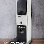 A white restaurant kiosk touchscreen stands against a gray wall on a wooden floor. The kiosk features a screen, a slot, and the word “KIOSK” with a logo on its lower section. “KIOSK” is overlaid in large text at the bottom left.