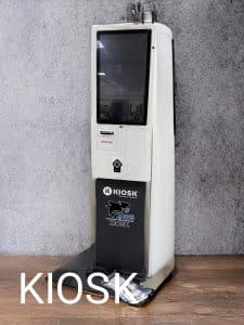 KIOSK Information Systems kiosk with TPGi JAWS -- A white restaurant kiosk touchscreen stands against a gray wall on a wooden floor. The kiosk features a screen, a slot, and the word “KIOSK” with a logo on its lower section. “KIOSK” is overlaid in large text at the bottom left.