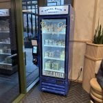 Smart Food Fridgevending machine from Royal Blue Grocery, reminiscent of modern kiosks endorsed by the National Restaurant Association, is stocked with various items. Situated near a glass door in a tiled room, a person is partially visible seated beside the machine.