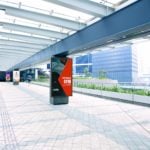 A modern, open-air covered walkway with tiled flooring leads through an urban area. A digital kiosk, a recent winner of digital signage awards, displays an ad for a fitness gym. Green plants line the walkway on the left, and tall buildings are visible in the sunny background.