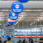 The airport terminal features blue and red signs for Deltas self-tagged bag drop and Sky Priority. Passengers queue under modern bright lighting, while TSA and CLEAR signage is visible. Digital signage enhances the area’s efficiency, guiding travelers to multiple check-in counters.