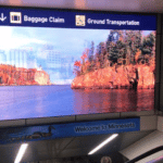 A large piece of airport digital signage showcases a scenic view of a lake with autumn-colored trees. Signs for baggage claim and ground transportation hover above, with a welcoming Welcome to Minnesota message just beneath the vibrant display.