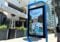 A digital display, showcasing the best kiosk design, features an ad of a smiling person in a helmet with the text Learn about city programs. The backdrop includes the iconic Portofino Plaza building and a passerby strolling along.