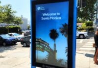 A digital sign on a city sidewalk, exemplifying the best kiosk design, warmly displays Welcome to Santa Monica. In the background, palm trees sway near signs for a fishing and boating café. Cars, a traffic light, and a pedestrian crossing complete the scene under a clear blue sky.