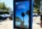 A digital sign on a city sidewalk, exemplifying the best kiosk design, warmly displays Welcome to Santa Monica. In the background, palm trees sway near signs for a fishing and boating café. Cars, a traffic light, and a pedestrian crossing complete the scene under a clear blue sky.
