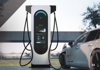 A modern EV charging station with a sleek design and digital display is connected to a white car. Situated outdoors with trees in the background, this station, akin to vital healthcare for vehicles, features multiple charging cables for efficient energy replenishment.