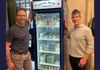 Two people stand beside a blue display fridge labeled Grab & Go from Royal Blue Grocery. Resembling a Smart Food Vending Kiosk, its stocked with packaged foods and beverages. They are indoors, near a window with a plant adding to the cozy ambiance.