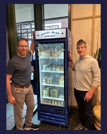 Two people stand beside a blue display fridge labeled Grab & Go from Royal Blue Grocery. Resembling a Smart Food Vending Kiosk, its stocked with packaged foods and beverages. They are indoors, near a window with a plant adding to the cozy ambiance.