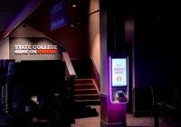 Inside a Dunkin coffee shop, dimly lit with a bright self-service kiosk reminiscent of those in Europe on the right. A sign reads STATE COLLEGE RUNS ON DUNKIN near stairs to the left. A PICK UP sign hangs above.