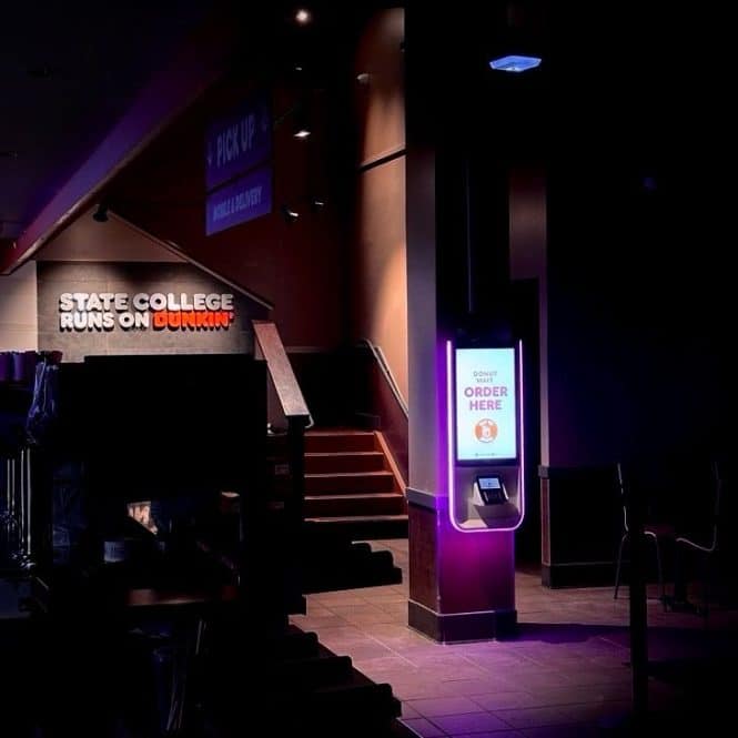 Inside a Dunkin coffee shop, dimly lit with a bright self-service kiosk reminiscent of those in Europe on the right. A sign reads STATE COLLEGE RUNS ON DUNKIN near stairs to the left. A PICK UP sign hangs above.