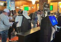 People use AI-powered self-check-in kiosks at a hotel lobby. The kiosks have screens displaying options. The background shows a sign for essentials and hotel services. The setting has warm lighting and a carpeted floor, enhancing the efficient, tech-savvy ambiance.