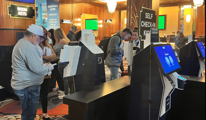 People use AI-powered self-check-in kiosks at a hotel lobby. The kiosks have screens displaying options. The background shows a sign for essentials and hotel services. The setting has warm lighting and a carpeted floor, enhancing the efficient, tech-savvy ambiance.