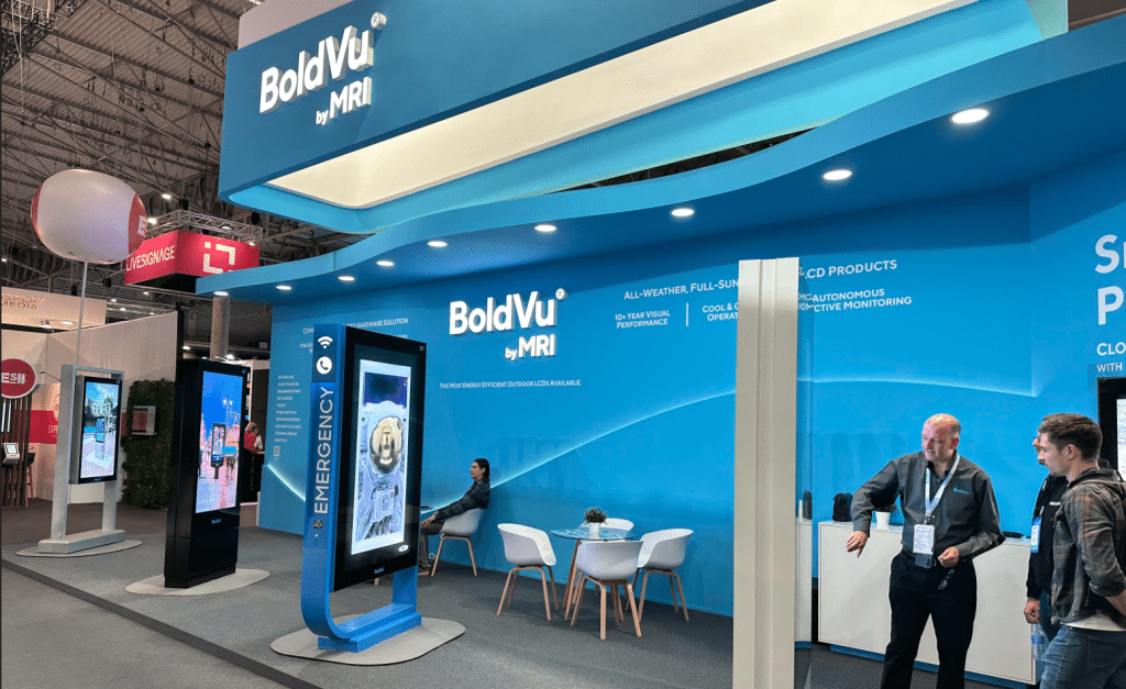A trade show booth featuring BoldVu by MRI offers an interactive experience. The large digital display highlights The Emergency as two people converse nearby. With a blue, curved design, the booth exudes a sleek infocomm vibe, complete with branding and seating at the back.
