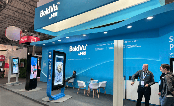 A trade show booth featuring BoldVu by MRI offers an interactive experience. The large digital display highlights The Emergency as two people converse nearby. With a blue, curved design, the booth exudes a sleek infocomm vibe, complete with branding and seating at the back.