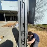 A person is crouched down, skillfully working on the wiring of a tall, metallic structure that is part of the Smart City initiative outdoors. The structure stands near the entrance of North Carolina’s Charles A Lyons Science Complex, with several boxes and shadows cast on the ground.