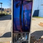 A tall outdoor digital display in a Smart City setting features a screensaver of a palm tree and sailboat. Its lower panel is open, revealing internal electronics. A person from Peerless-AV crouches beside it, working with a laptop to ensure seamless operation in North Carolinas vibrant environment.