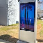 A Peerless-AV digital kiosk showcases a desktop wallpaper of a tropical beach scene, complete with palm trees and a sailboat. Situated in North Carolina, this smart city feature is positioned on a grass patch by the sidewalk, framed by nearby buildings and trees.