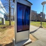 A digital kiosk from Peerless-AV with a large touchscreen display stands on a grassy area next to a sidewalk. It showcases a beach-themed desktop wallpaper. This Smart City feature enhances the outdoor scene under the clear sky, surrounded by North Carolinas buildings and trees.