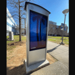 A Peerless-AV outdoor digital display panel, showcasing a computer desktop with palm trees, graces the sidewalk. Nestled near a modern building and lush trees under clear skies, it embodies the essence of a Smart City in North Carolina.