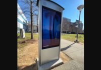 A Peerless-AV outdoor digital display panel, showcasing a computer desktop with palm trees, graces the sidewalk. Nestled near a modern building and lush trees under clear skies, it embodies the essence of a Smart City in North Carolina.