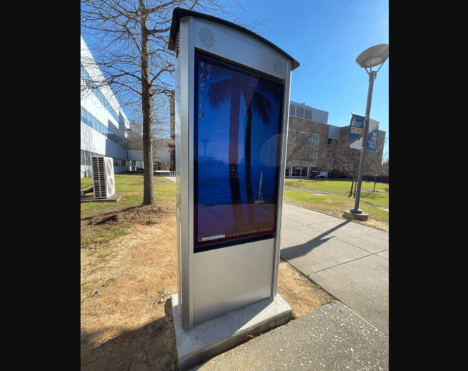 A Peerless-AV outdoor digital display panel, showcasing a computer desktop with palm trees, graces the sidewalk. Nestled near a modern building and lush trees under clear skies, it embodies the essence of a Smart City in North Carolina.