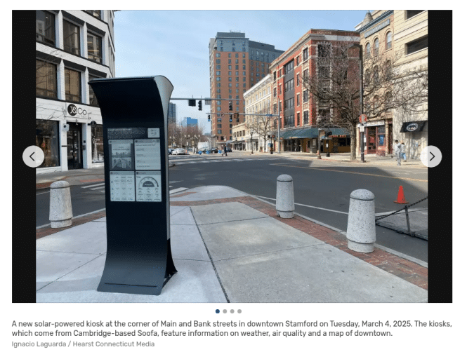 A solar-powered kiosk in downtown Stamford provides crucial wayfinding information amidst brick buildings and shops. The quiet street, free of pedestrians and vehicles, showcases the kiosks efficient design while offering real-time weather updates to passersby.