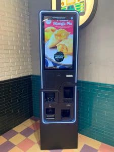 The McDonalds self-service kiosk features a vibrant touchscreen showcasing an ad for Mango Pie. With illuminated edges, it stands against a brick and tiled wall. Convenient payment options, including cash and coin, are available right below the screen.