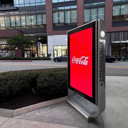 Boldvu by MRI digital signage wayfinding A digital emergency kiosk on a city sidewalk displays a red Coca-Cola advertisement. An office building, shops, parked cars, and pedestrians are visible in the background.