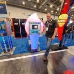 A man stands smiling beside a futuristic touchscreen kiosk at a carwash trade show tour, surrounded by colorful equipment displays and banners in a spacious exhibition hall.