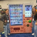 Two women stand smiling next to a vending machine filled with pet treats at a carwash trade show tour. The machine is decorated with colorful labels and images of animal snacks, featuring a digital display and branding on the front.