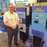 A man in a white shirt and green lanyard stands next to two blue vending machines labeled CHANGE and BILL BREAKER at a carwash trade show tour.