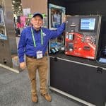 A man in a blue shirt and cap stands smiling next to a red vending or arcade machine at a carwash trade show. Wearing a badge, he is surrounded by other displays and equipment, ready to give visitors a tour of the booth area.