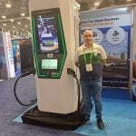 A person stands smiling next to a large electric vehicle charging station at an indoor expo, part of a carwash trade show tour, with informational banners and booths visible in the background.