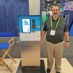 A man with glasses and a green lanyard stands next to a blue and white kiosk called The Wave at a carwash trade show. A wooden director’s chair is nearby in the booth, capturing the energy of this industry tour.