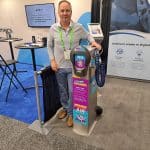 A man stands at a carwash trade show booth next to a digital air calibration machine for tires. The booth showcases banners and promotional materials about tire care, air calibration services, and details of their trade show tour.