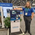 A man with long hair and a beard, wearing a blue polo and gray pants, stands next to a Hamilton Car Wash payment terminal during a carwash trade show tour. Bright overhead lights illuminate the scene.