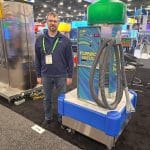 A man with glasses and a lanyard stands next to a colorful Turbo Vacuum machine during a carwash trade show tour, surrounded by various booths and machines in the background.